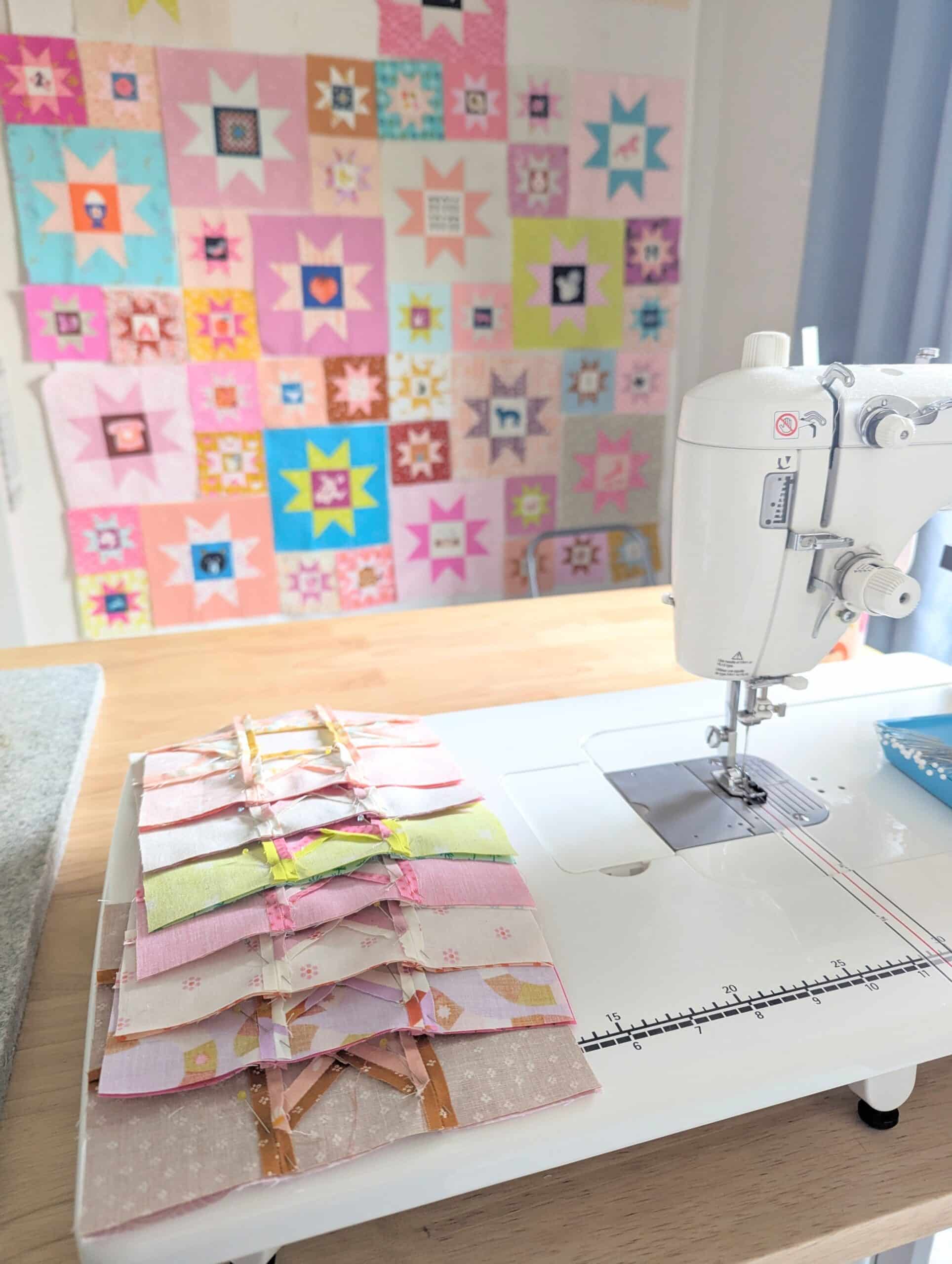 Quilt blocks lay on a sewing machine bed while colourful blocks can be seen on the wall in the background.