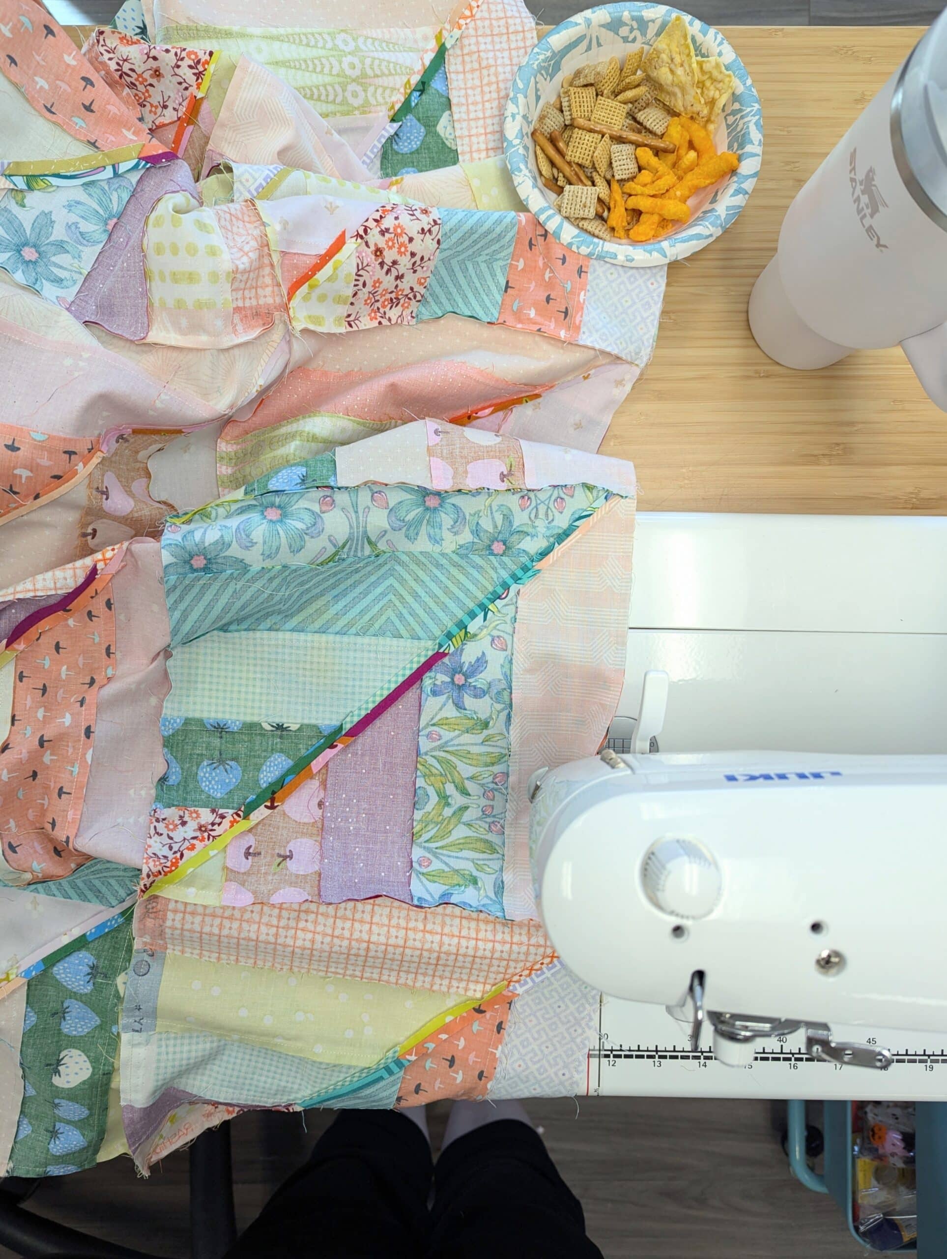 An overhead view of a sewing machine on a wooden table, with a bowl of snacks and a water bottle.