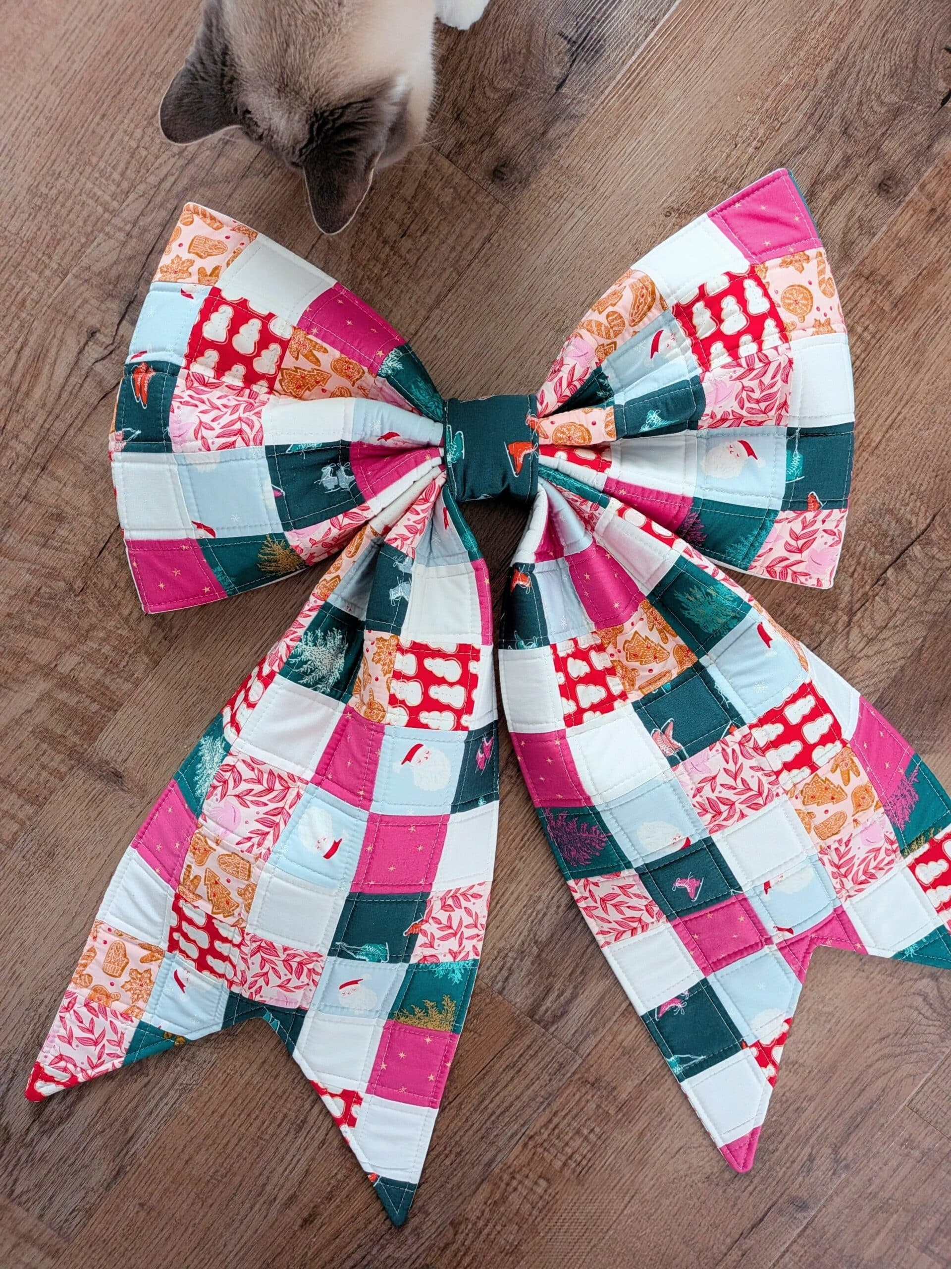 A patchwork bow lies on a wooden floor with a cat in the frame.