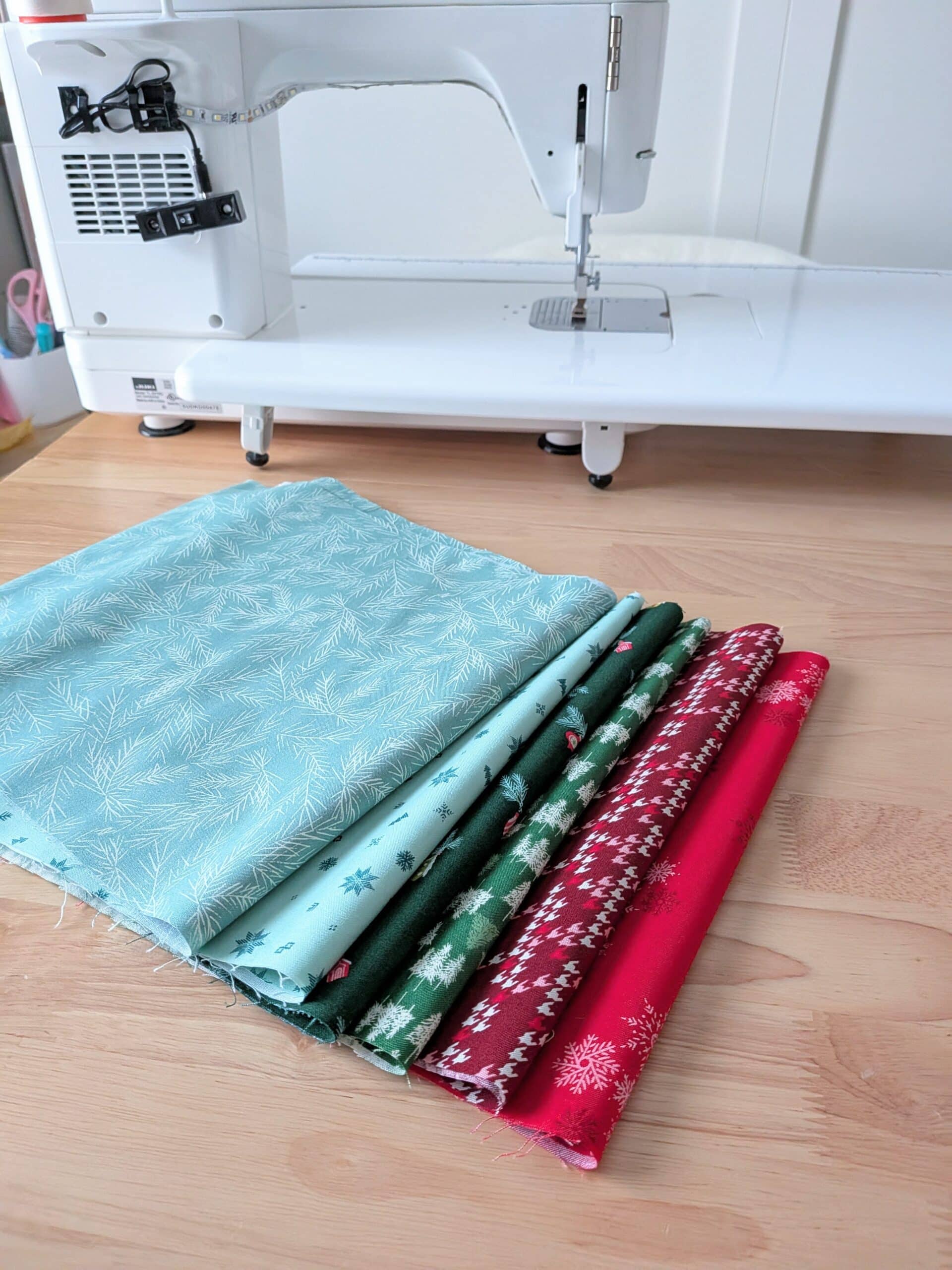 Five pieces of fabric (blue, red, and green) lay on a worktable with a sewing machine in the background.