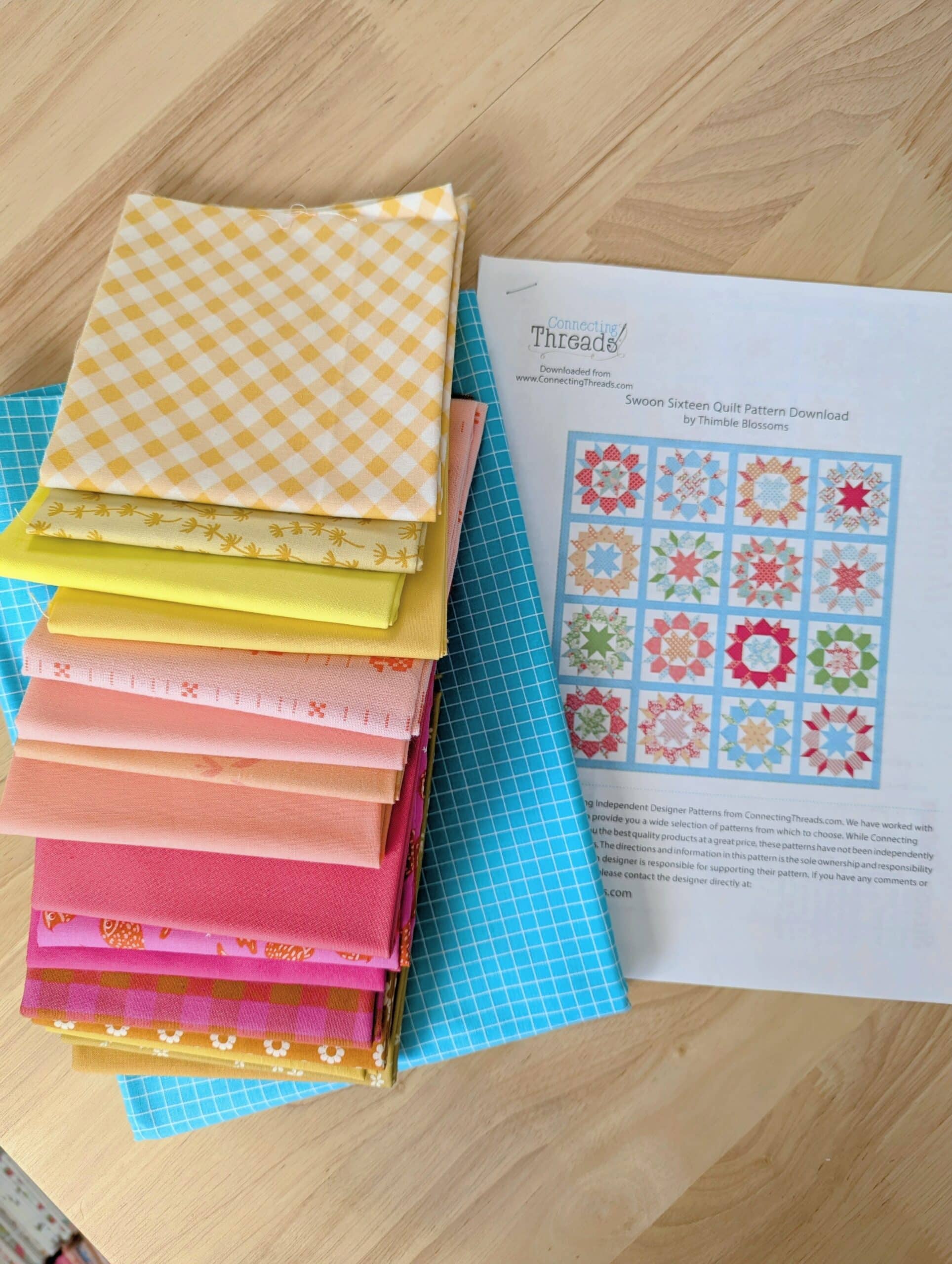 A stack of pink and yellow fabric lies on top of a larger piece of blue grid fabric next to a printed quilt pattern.
