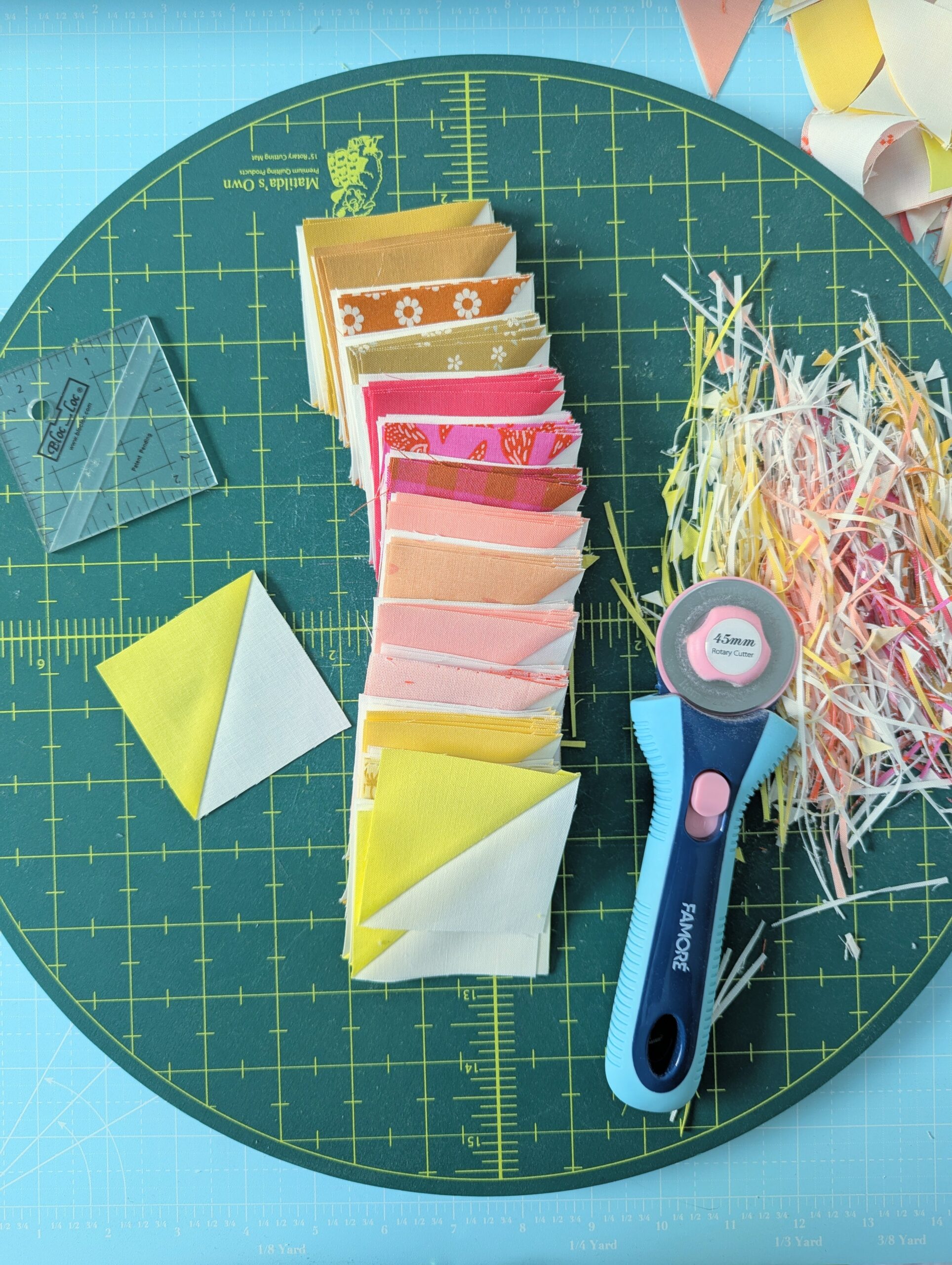 A stack of completed and trimmed half square triangle blocks sit on a rotating cutting mat along with a small Bloc Loc Ruler and rotary cutter.