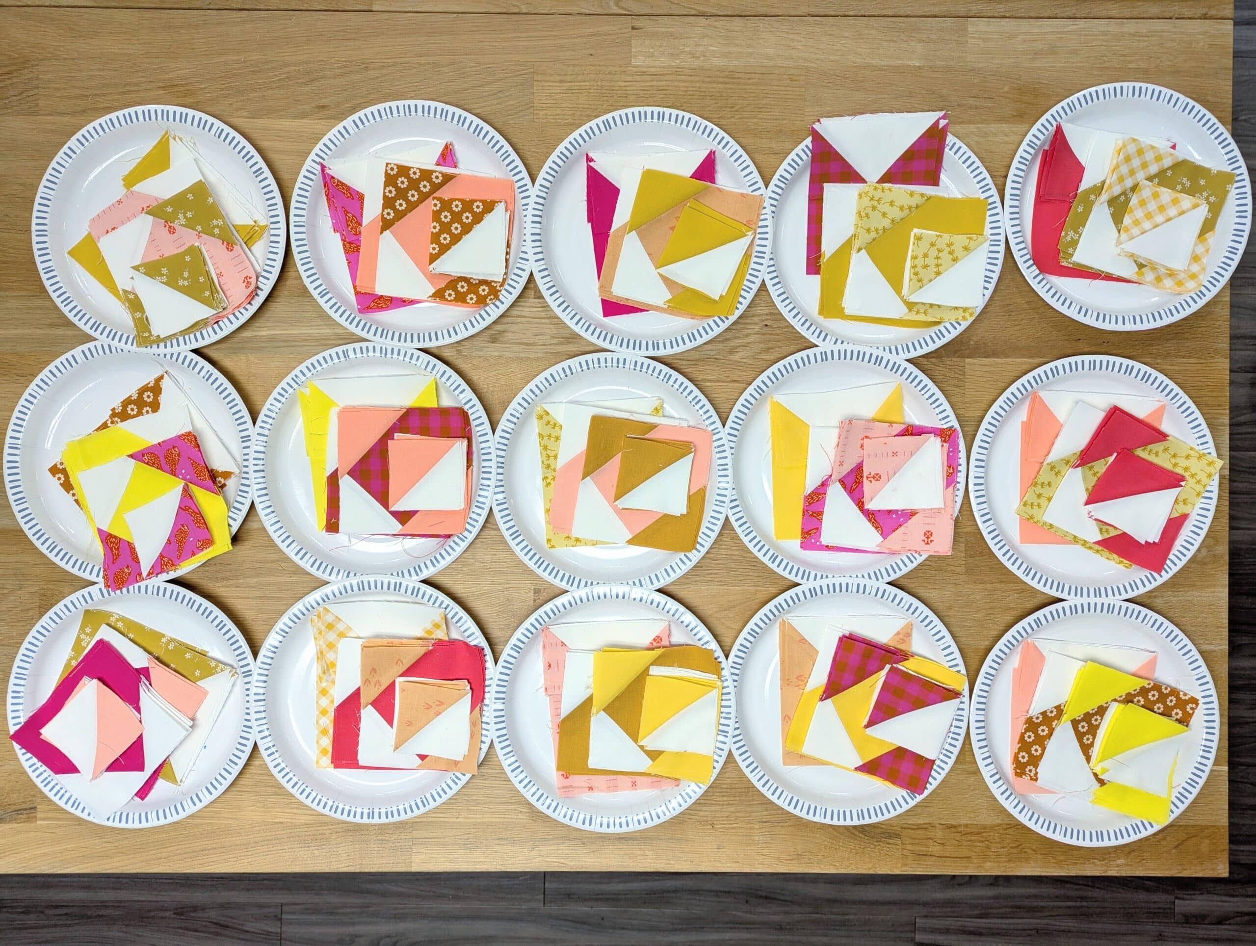 Fifteen paper plates are arranged on a wooden desk, each one with a stack of colourful pieces of fabric.