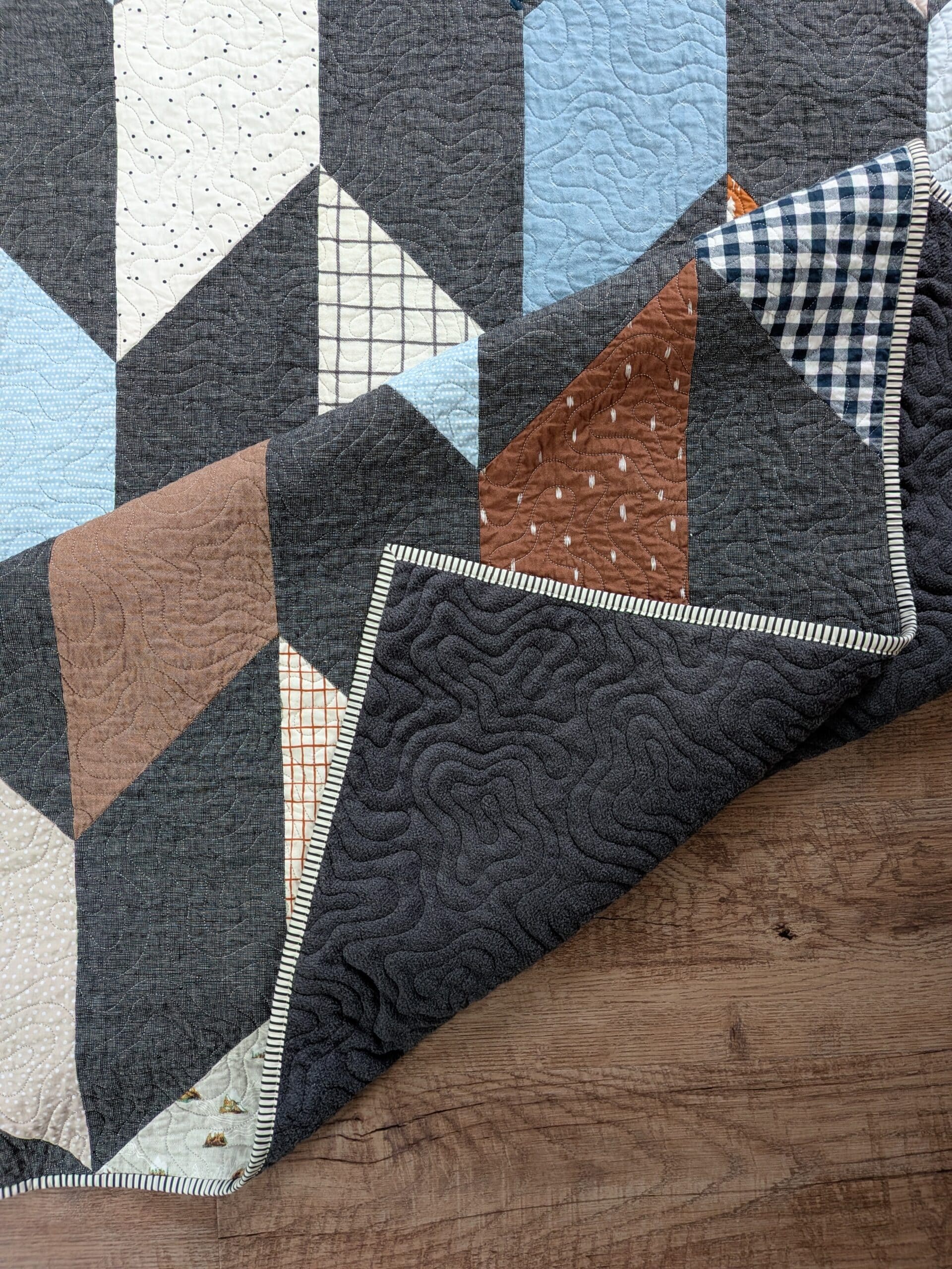 A chevron quilt lays on a brown wood floor, with one corner flipped onto itself twice to reveal a fuzzy dark gray backing fabric.