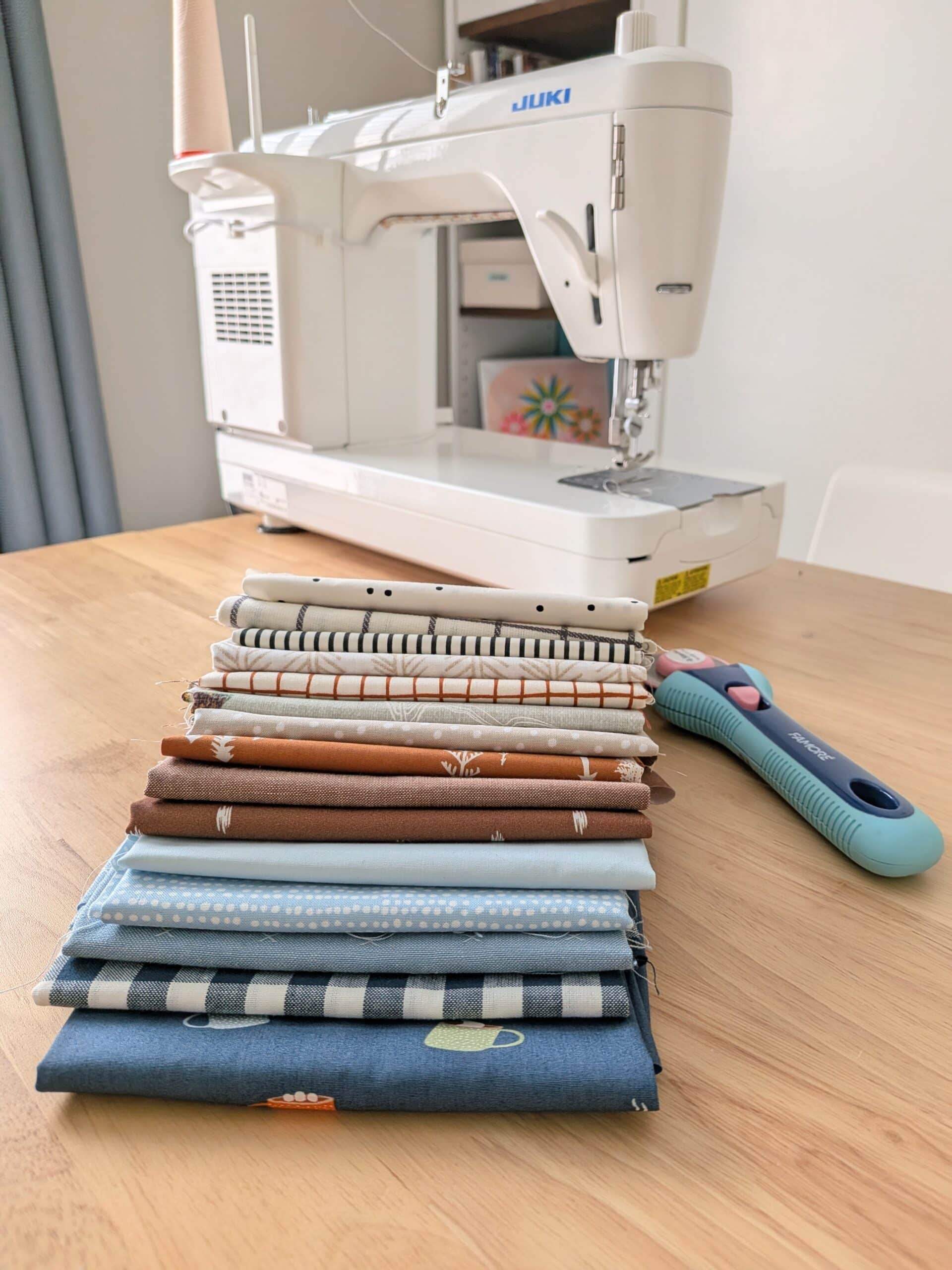 A stack of 15 pieces of fabric are fanned out on a wooden table with a sewing machine in the background.