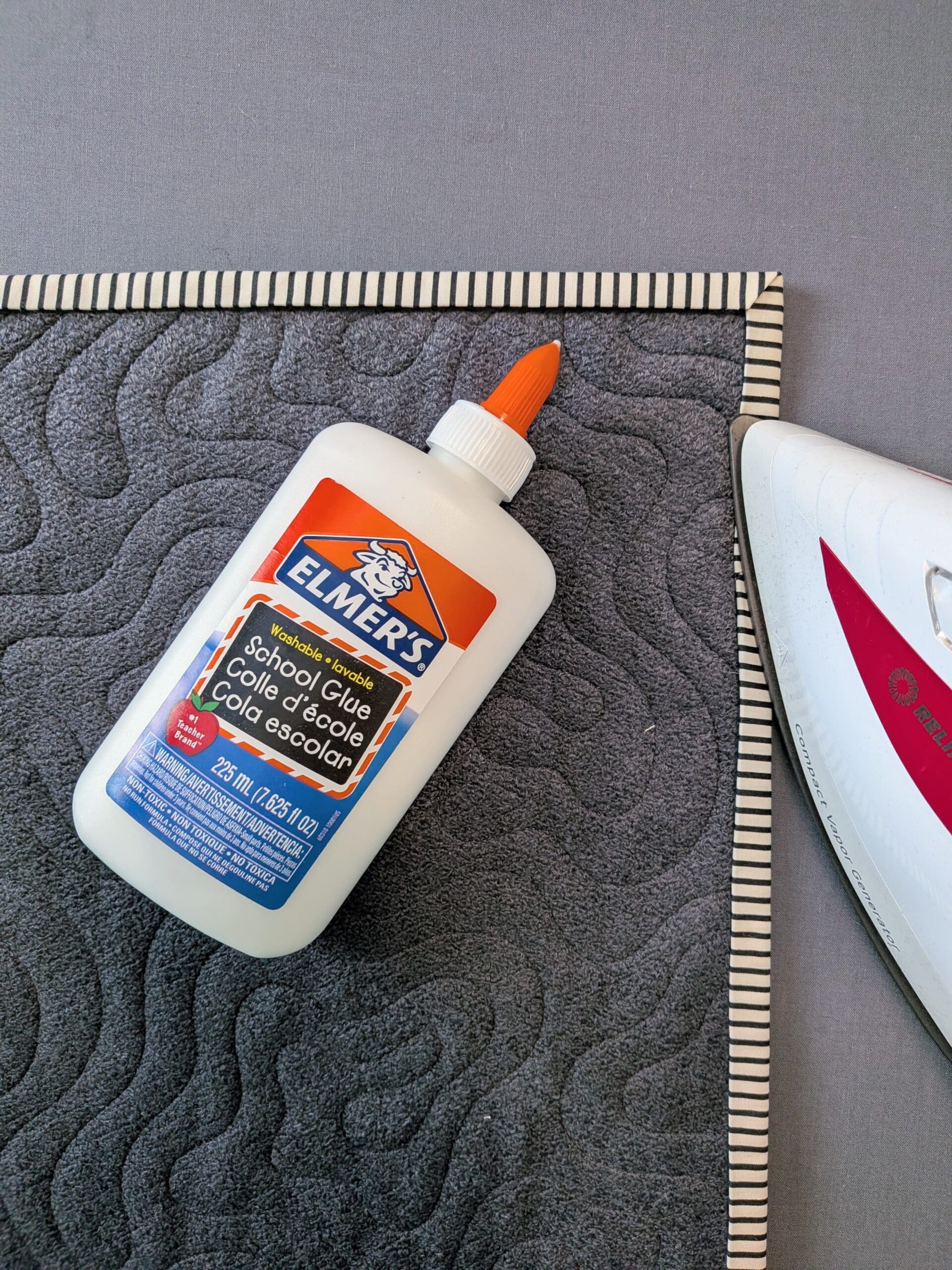 An iron presses the striped binding fabric near one corner of a quilt. A bottle of Elmer's School Glue lays on top of the quilt.