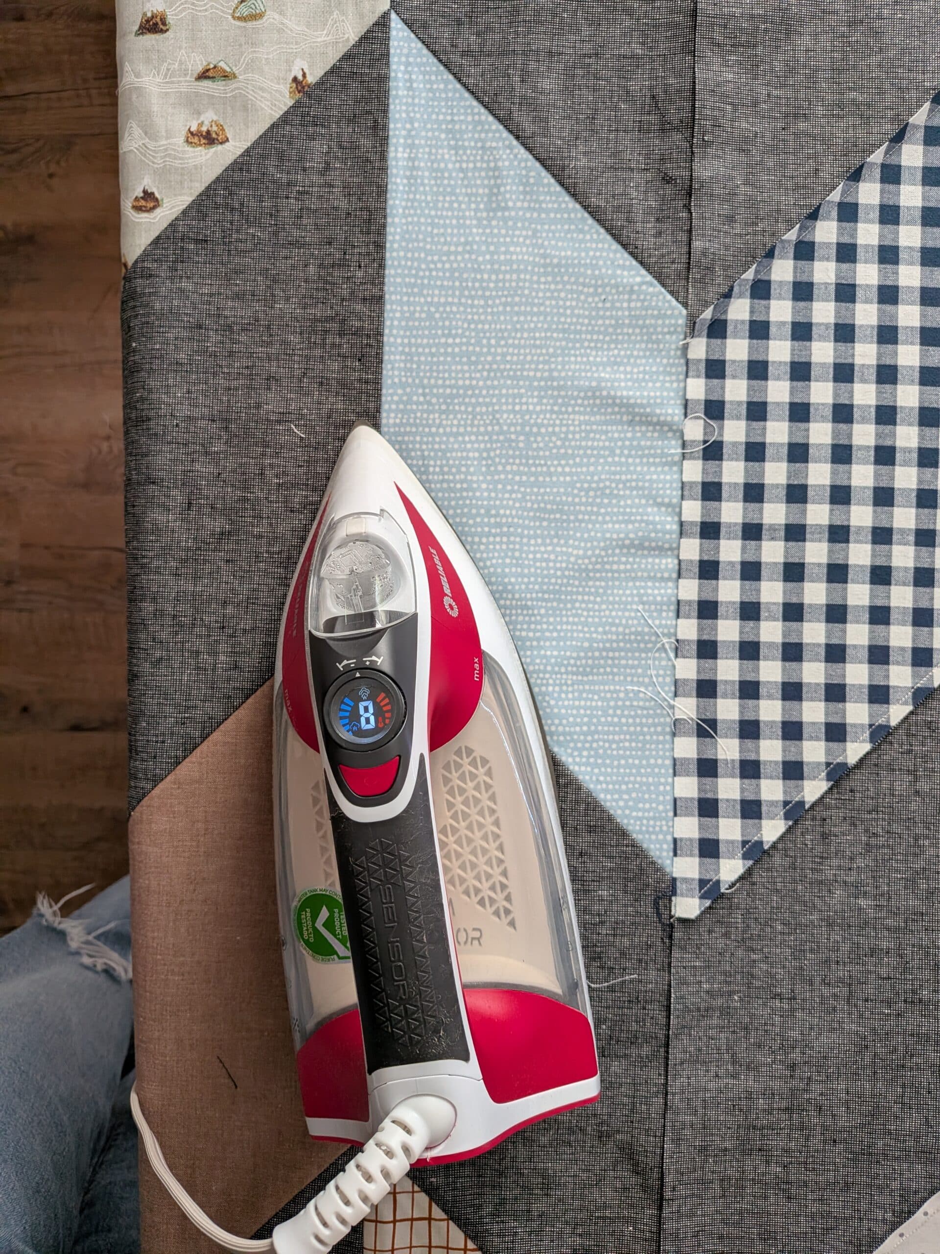 A red iron lays on top of a quilt top on an ironing board.