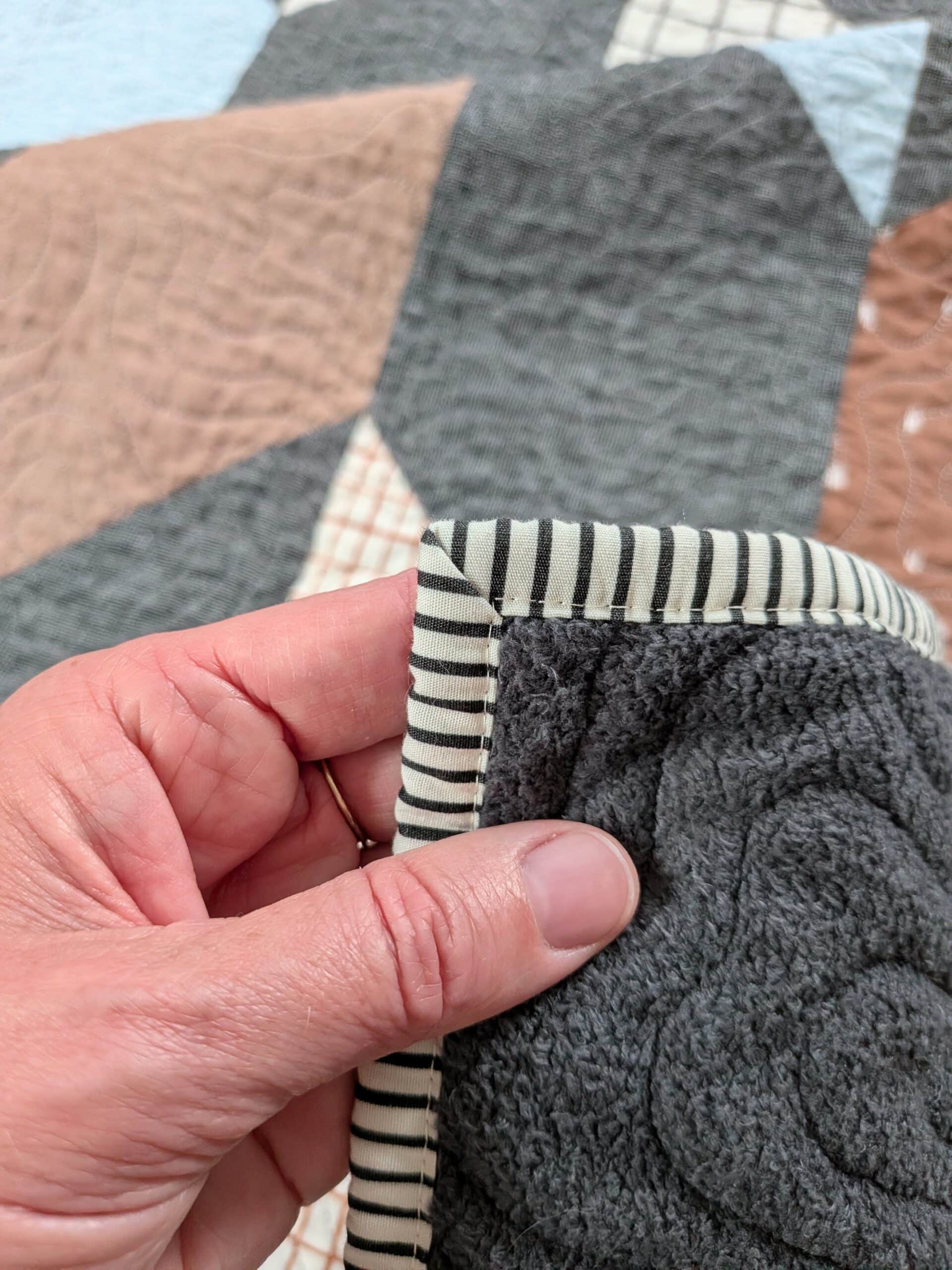 Shelly holds one corner of a quilt, showing the striped binding to be attached with a line of stitches on the back of the quilt.