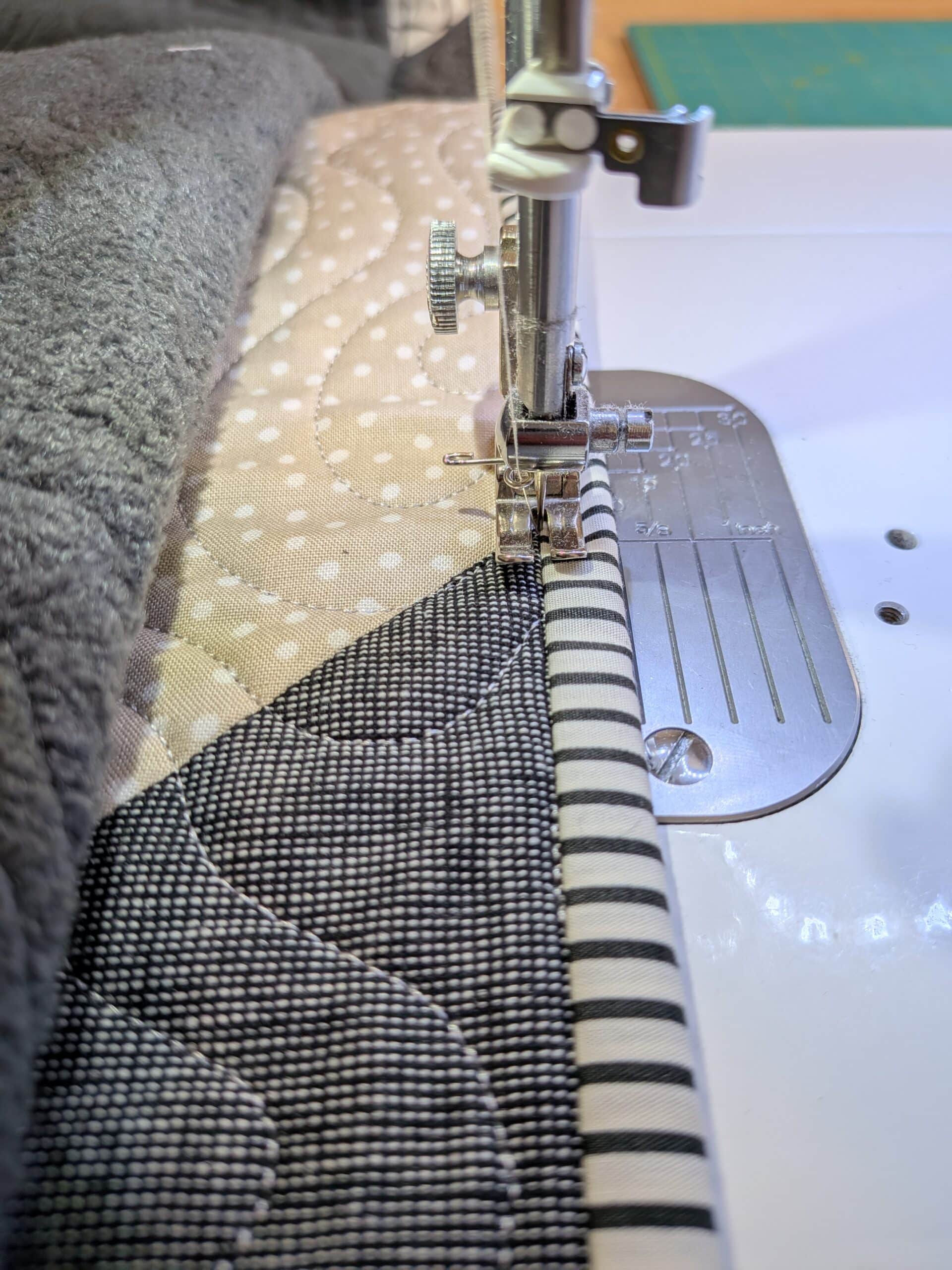 The needle of a sewing machine falls between the striped binding fabric and quilt top.