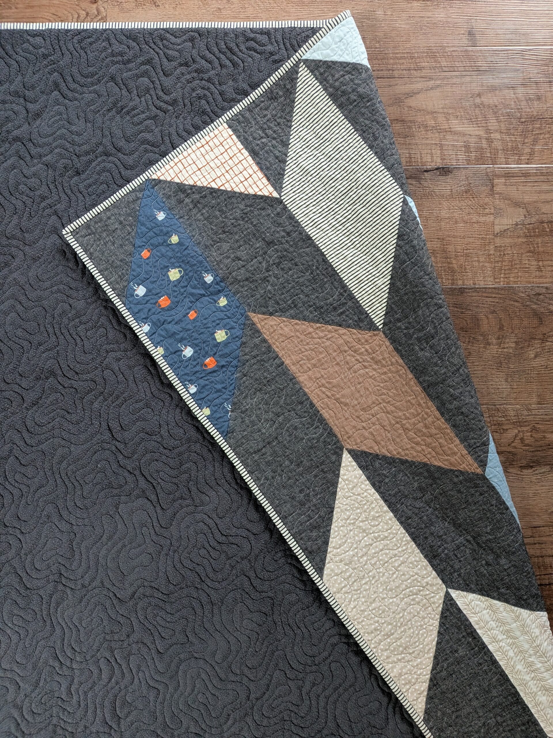 A quilt with a fuzzy dark gray backing lays face down on a brown wooden floor. One corner of the quilt has been flipped over to reveal a chevron patchwork pattern.